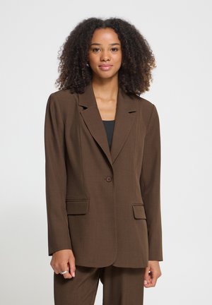 Donna con capelli ricci che indossa una giacca e pantaloni marroni abbinati, in piedi davanti a uno sfondo chiaro e uniforme.