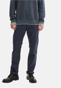 Navy modre, slim-fit hlače z gladko teksturo. V kombinaciji z temnim puloverjem, ki ima svetlo obrobo, in črnimi čevlji na vezalke.