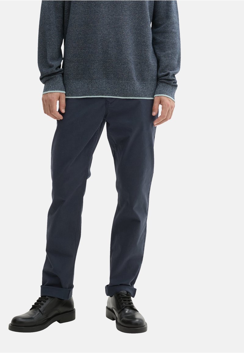 Navy modre, slim-fit hlače z gladko teksturo. V kombinaciji z temnim puloverjem, ki ima svetlo obrobo, in črnimi čevlji na vezalke.