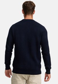 Marineblå strikket genser med et strukturert chevronmønster, rund hals og ribbestrikkede ermer og kant. Stoffet virker varmt og slitesterkt.