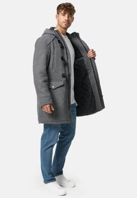 Cappotto grigio in misto lana con chiusure a fermaglio, grandi tasche, cappuccio e fodera trapuntata nera. Indossato sopra una camicia bianca e jeans.