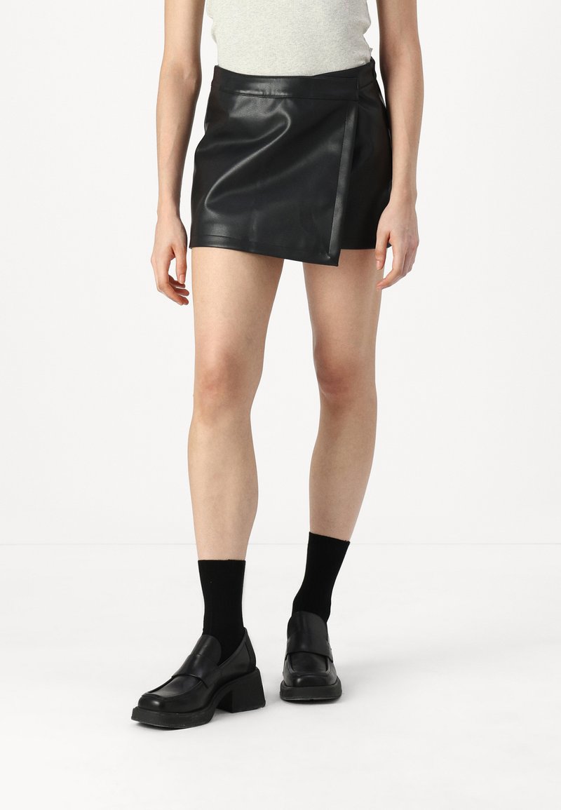 Falda corta de cuero negro con una textura suave, cinturilla alta y longitud mini. Combinada con unos mocasines negros gruesos y calcetines negros.