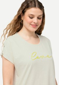 Camiseta de manga corta color beige claro, hecha de un tejido suave, con texto amarillo en cursiva que dice "amor" y detalles de botones decorativos en los hombros.