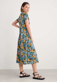Blumenkleid mit kurzen Ärmeln, knielang, in Schwarz mit teal-, orange- und lila-blumen, kombiniert mit schwarzen Sandalen mit Riemen.
