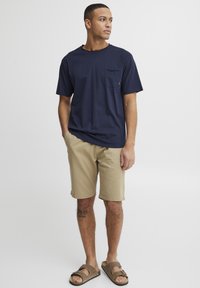 Tricou bleu marin cu maneci scurte și buzunar frontal, asortat cu pantaloni scurți bej până la genunchi și sandale maro tip papuci. Design simplu și casual.