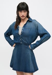 Rifľové šaty v tmavomodrej farbe s gombíkovým zapínaním vpredu a elastickým pásom. Majú dlhé rukávy a vyšívané logo "Diesel" na hrudi.