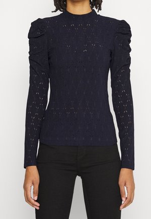 Person iført en tætsiddende, langærmet navy bluse i blonde med oppustede skuldre og sorte bukser mod en ensfarvet lys baggrund.