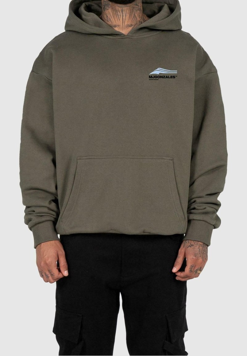 Homme portant un sweat à capuche vert olive avec poche avant et pantalon noir, tatouages visibles sur le cou et les mains, debout devant un fond uni.