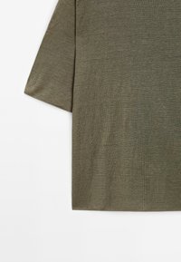 Oliwkowa koszula z krótkim rękawem o teksturowanej, miękkiej tkaninie. Design charakteryzuje się luźnym krojem i prostymi, czystymi liniami bez ozdób.