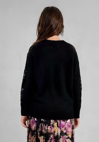 Maglione lavorato a maglia nero con scollo rotondo e orlo a costine, caratterizzato da dettagli in pizzo alle maniche. Abbinato a una gonna floreale plissettata.