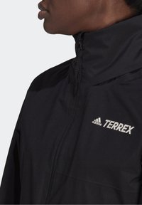 Svart Terrex-jacka i slät tyg, med hög krage, frontdragkedja och vit Adidas-logotyp på vänster bröst.