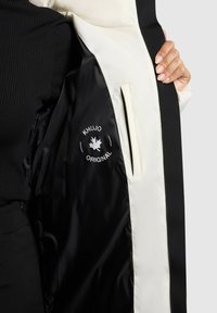 Interior de chaqueta negra y crema con textura suave, que presenta un logo y un bolsillo con cremallera en el lado izquierdo.