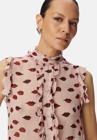 Roze mouwloze blouse met ruches aan de hals en schouders, versierd met rode lipafdrukken. Gemaakt van doorzichtig materiaal.