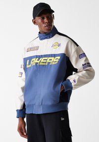 Lakers jacket featuring a color block design in blue, cream, and black, with prominent logo, embroidered patches, and ribbed cuffs.