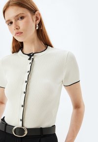 Rippiertes Kurzarmoberteil in Creme mit schwarzem Besatz, mit runder Ausschnitt, Knopfverschluss und einer Schleifenakzent am Kragen.