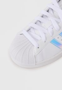 Bijela tenisica s teksturiranim plastičnim vrhom, ravnim bijelim vezicama i iridescentnim trakama sa strane. Glatki sintetički materijal.