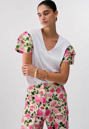 Žena tamne kose u bijeloj majici s V-izrezom i cvjetnim puf rukavima te hlačama u istom uzorku, nosi zlatne narukvice i naušnice, gleda prema dolje.