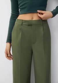 Olive green tailored trousers with a smooth, textured fabric, featuring front pleats and a flat waistband. No visible hardware or patterns.