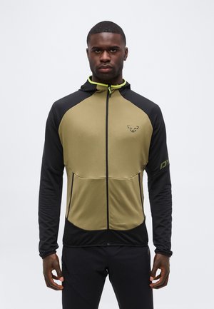 Bărbat care poartă o hanorac cu fermoar negru și măsliniu cu glugă, stând cu fața spre înainte pe un fond alb simplu.