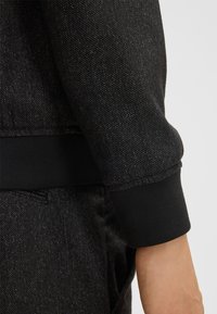 Tessuto di denim nero con una leggera trama, dotato di un polsino nero a coste che offre una vestibilità aderente. Nessun motivo o accento visibile.