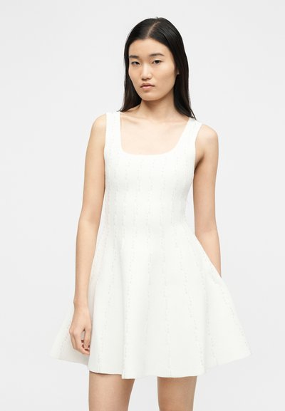 Νεαρή γυναίκα με ίσια μαύρα μαλλιά, ντυμένη με λευκό πλεκτό αμάνικο φόρεμα με τετράγωνη λαιμόκοψη και φαρδύ κάτω μέρος.