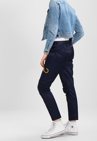 Osoba w ciemnoniebieskich spodniach z żółtą grafiką na udzie, skrojonym jasnoniebieskim dżinsowym kurtce i białych trampkach na wysokiej podeszwie.