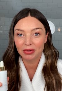 Bouteille blanche avec pipette, capuchon doré, tenue devant un fond en carrelage. Le sujet a de longs cheveux bruns, porte une robe blanche. Peau claire et maquillage léger.