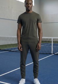 Mees, kes kannab tumerohelist sportlikku riietust ja valgeid tenniseid, seisab sinisel sise-tenniseväljakul võrgu lähedal.