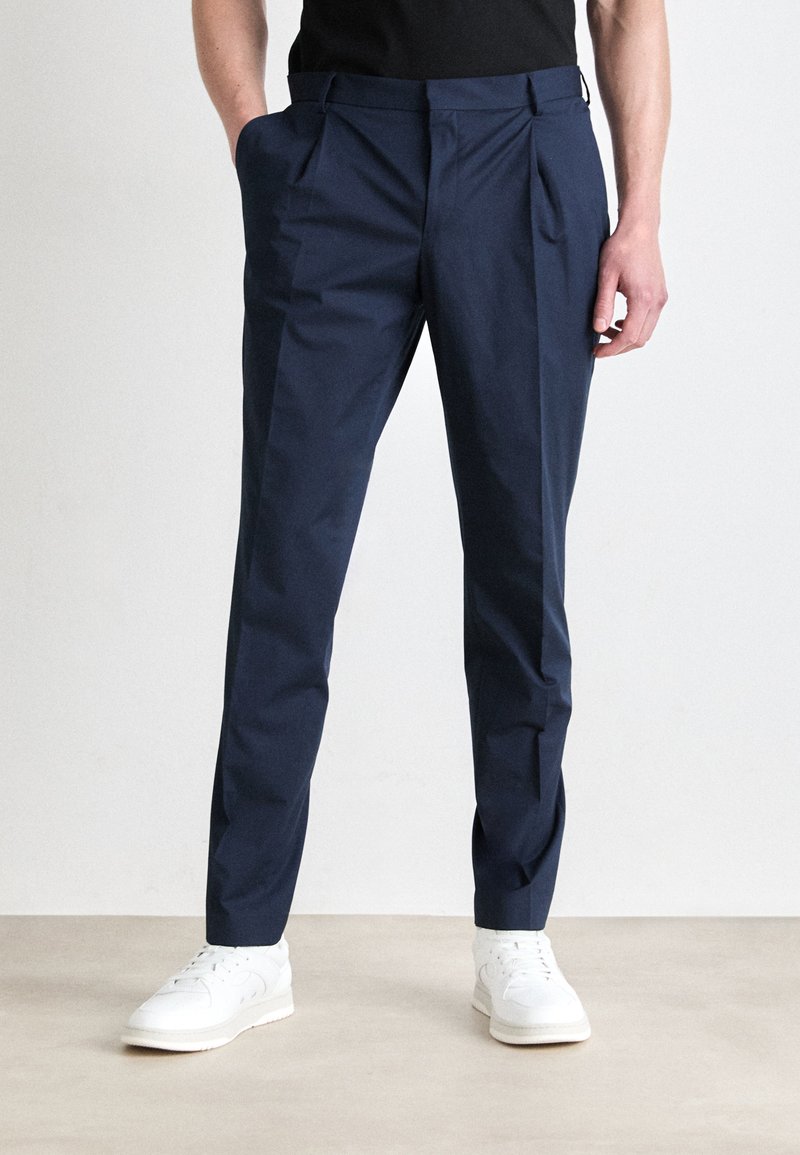 Navy blue trousers with a tailored fit, featuring pleats at the front, side pockets, and a smooth fabric texture. Worn with white sneakers.