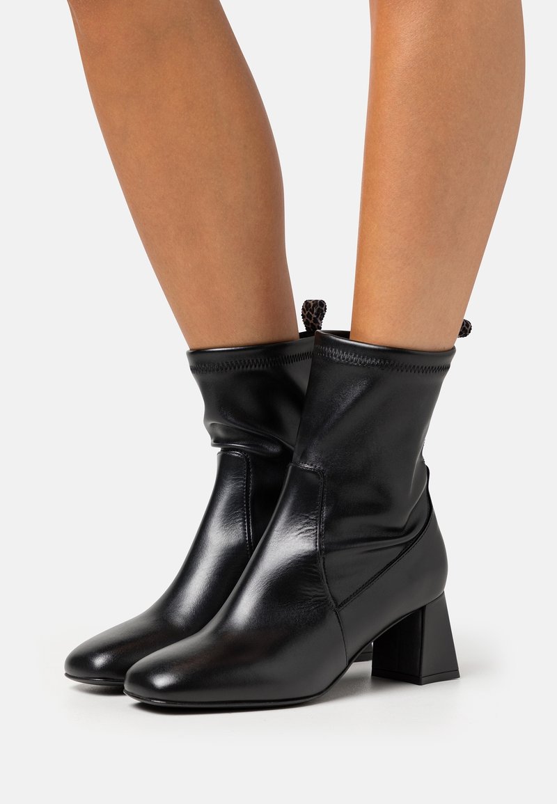 Une personne porte des bottines noires brillantes à talons carrés et bout arrondi, debout sur un fond clair.