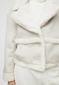 Chaqueta de imitación de piel de color crema con forro de piel sintética, cuello, dos bolsillos horizontales y dos botones. Exterior liso con puños y dobladillo suaves y texturizados.
