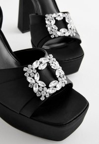 Black platform sandals with a smooth satin texture, featuring a rhinestone-embellished buckle and a chunky heel design.