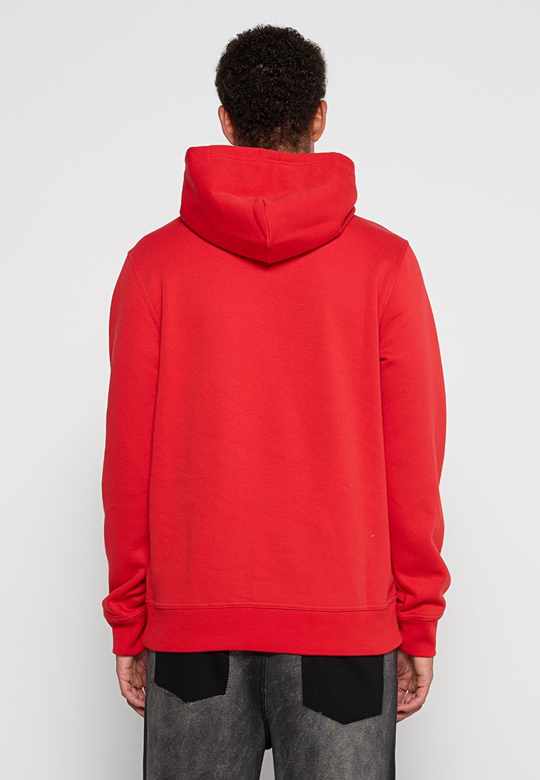 Persona con el cabello rizado corto que lleva una sudadera roja simple vista desde atrás, combinada con pantalones negros y grises.