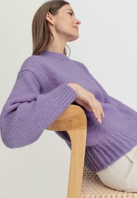 Violetti neulottu neulepaita, jossa on ribattu helma ja hihansuukki, henkilö istuu puutuolilla. Pehmeä rakenne ja rento istuvuus.