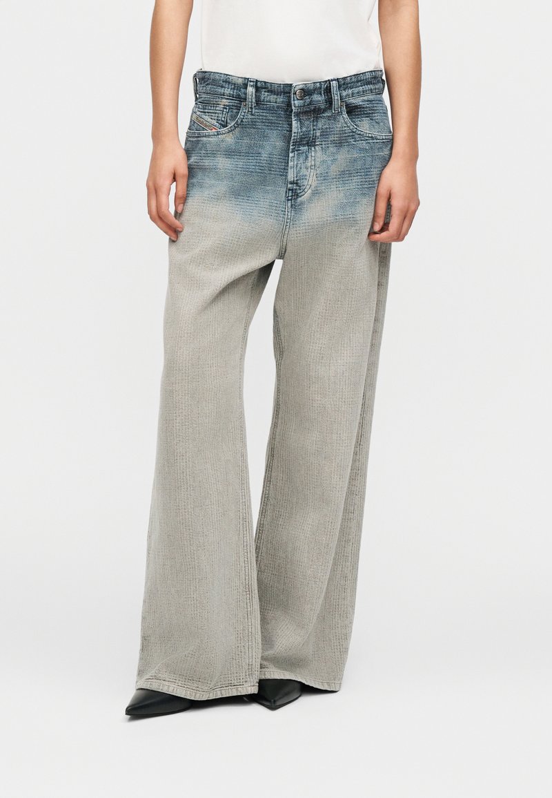 Κάτω μισό μέρος ενός ατόμου που φοράει ψηλόμεσα, φαρδιά, ξεβαμμένα τζιν παντελόνια και μαύρα μυτερά παπούτσια, στέκεται σε απλό φόντο.