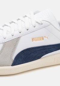 Vita sneakers med grå mocka och marinblå accenter. Har flata snören, strukturerad sula och en gyllene Puma-logotyp på sidan.