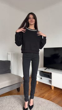 Felpa nera con decorazioni sul collo, abbinata a jeans skinny neri e scarpe con tacchi neri. Sfondo e mobili semplici.