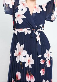 Robe plissée bleu marine à grand imprimé floral blanc et rose, col en V, manches courtes transparentes et ceinture nouée à la taille.
