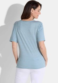 Helles blau gestreiftes T-Shirt mit kurzen Ärmeln, rundem Halsausschnitt und weichem Material. Getragen mit weißen Hosen, von hinten betrachtet.