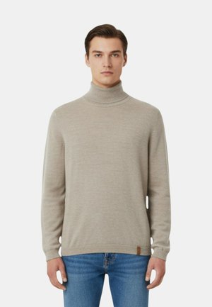 Giovane uomo con indosso un maglione beige a collo alto e jeans blu, in piedi contro uno sfondo semplice di colore grigio chiaro.