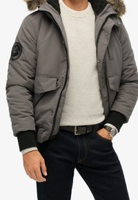 Grijze donsjas met een bontkraag, ritssluiting aan de voorkant en twee zakken vooraan. Draagt over een lichte trui, gecombineerd met donkere spijkerbroek en een horloge.