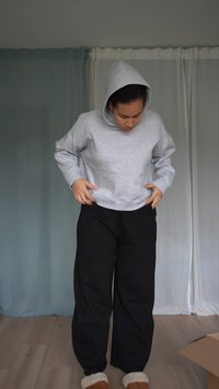 Szürke, kapucnis cropped hoodie, lazán megkötött fekete joggingnadrággal párosítva. A modell beltérben áll, világos színű fapadlóval és texturált háttérrel.