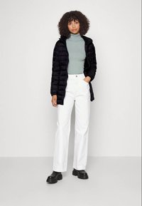 Casaco puffer preto, suéter de gola alta verde com textura, calças brancas de perna larga e botins pretos robustos. Modelo de pé com as mãos nos bolsos.