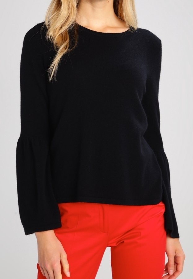 Femme portant un haut noir à manches longues avec des poignets évasés et un pantalon rouge vif, debout devant un fond clair uni.