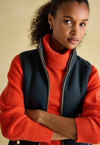 Námornícky fleecový vestu cez červený hrubý pletený rolák. Vesta má zips a bočné vrecká; rukávy svetra sú rebrované s textúrovanými vzormi.