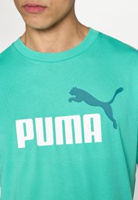 Άνδρας που φοράει πουκάμισο Puma τιρκουάζ με μεγάλο λευκό κεί μενο Puma και σκούρο τιρκουάζ λογότυπο πηδούντος πούμα στο μπροστινό μέρος.