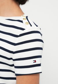 Κοντινή λήψη ενός λευκού πουκαμίσου με μαύρες οριζόντιες ρίγες, χρυσά κουμπιά στον ώμο και ένα μικρό κόκκινο, λευκό και μπλε λογότυπο στο μανίκι.