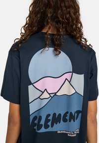 Marineblauwe t-shirt met een veelkleurige afbeelding van bergen en een cirkel, met roze, lichtblauwe en witte elementen.