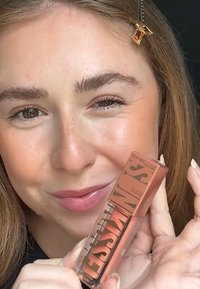 Maybelline flytande rouge i en genomskinlig tub med en persikoton, med en transparent botten och ett metalliskt rosafärgat lock med fet svart text.