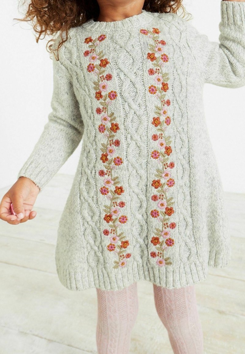 Enfant portant une robe-pull tricotée gris clair avec broderie florale verticale et collants roses à motifs.